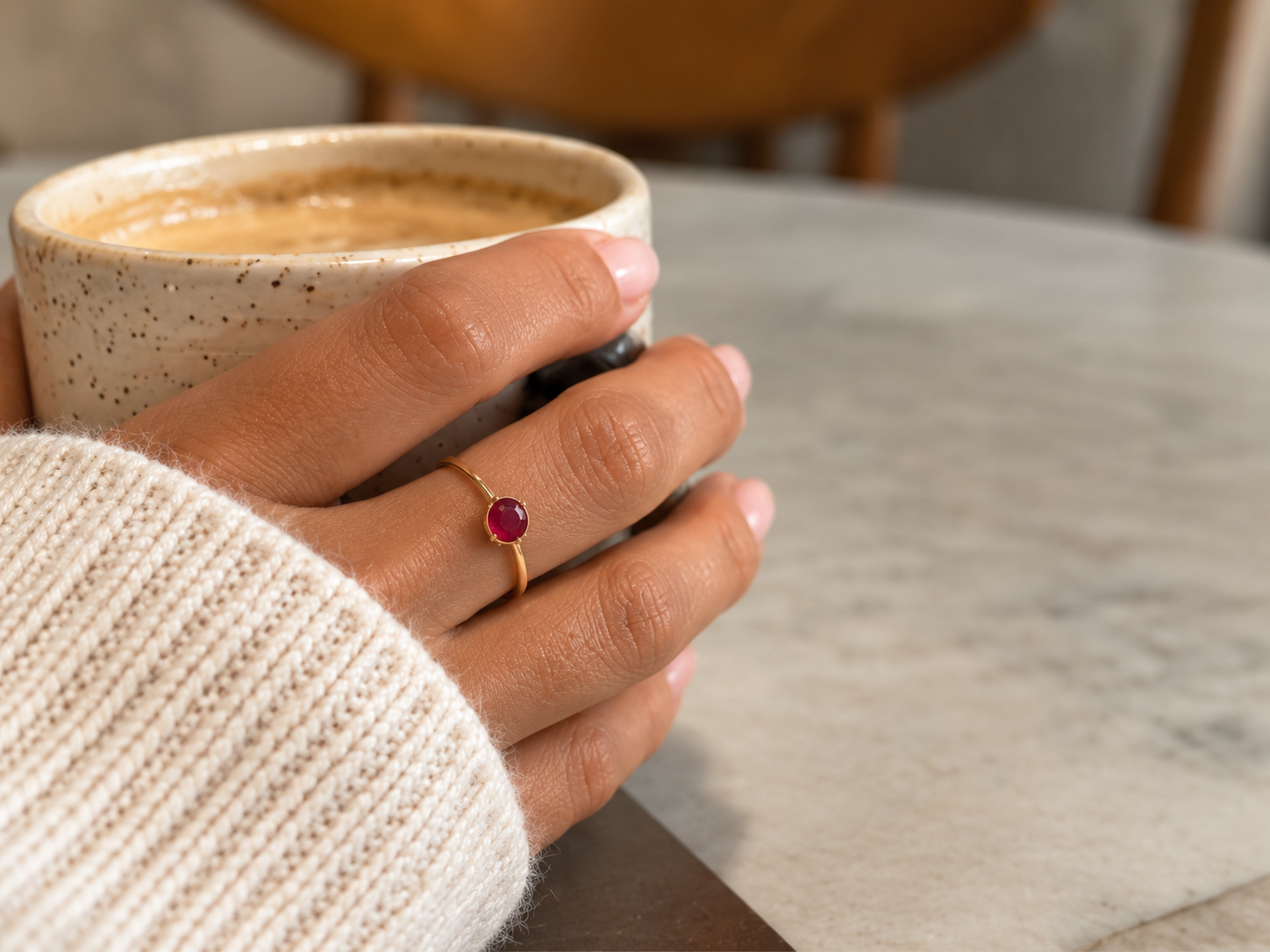 Hand with a gold ruby birthstone ring wrapped around a speckled coffee cup