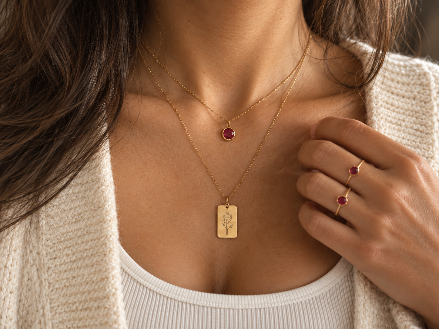 Layered gold necklaces with a ruby pendant and a birthstone flower charm, with matching ruby rings